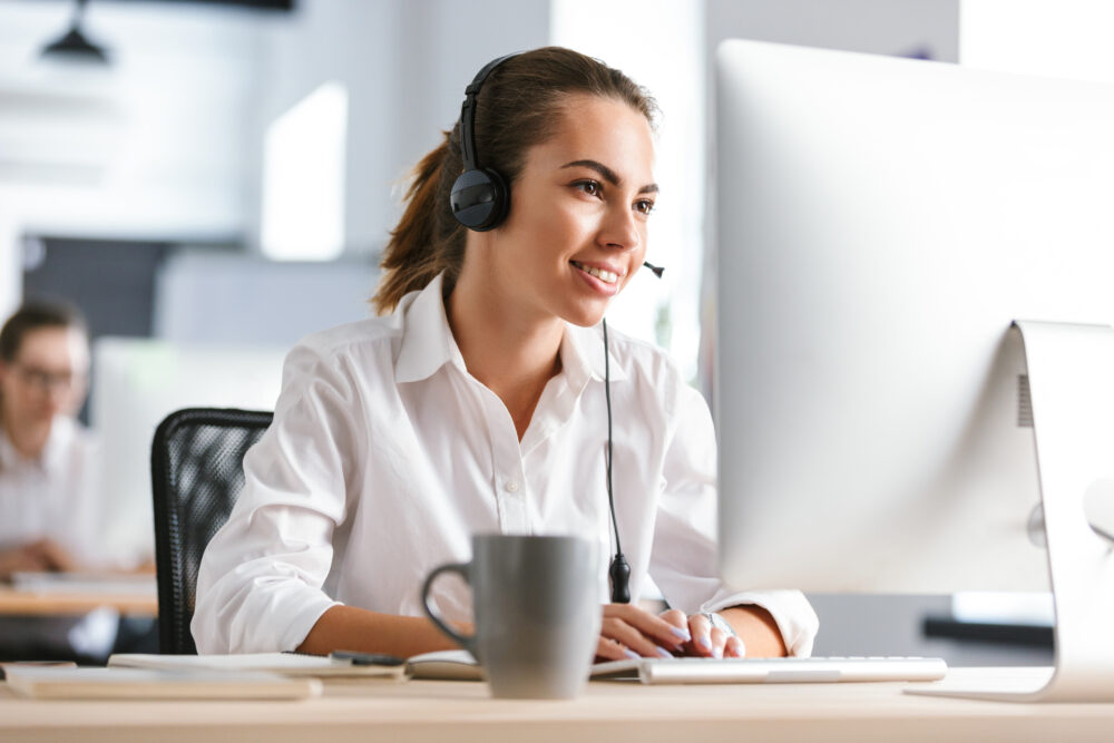 Happy cheerful business woman in office callcenter working with computer wearing headphones.