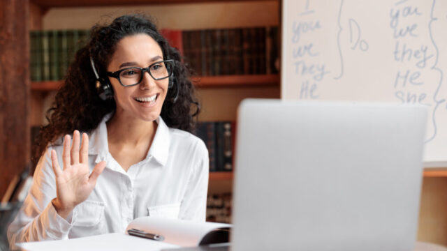Distance Education. Positive lady wearing glasses and wireless headest at virtual meeting, sitting at desk, having video call on laptop, waving to webcam. Woman studying or teaching online at home